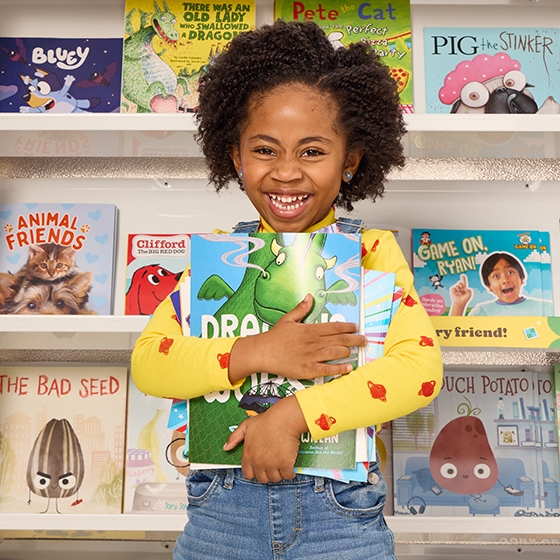 A small child smiles in front of a Book case holding several books in her arms
