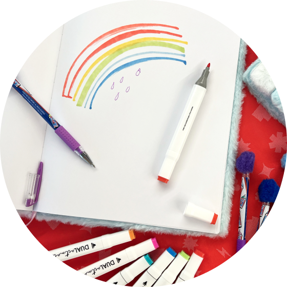 A notebook open to a drawing of a rainbow surrounded by markers and pens