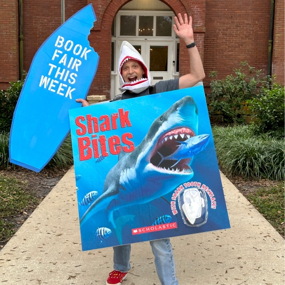 Man in Shark Bites book costume holds a sign saying Book Fair this week