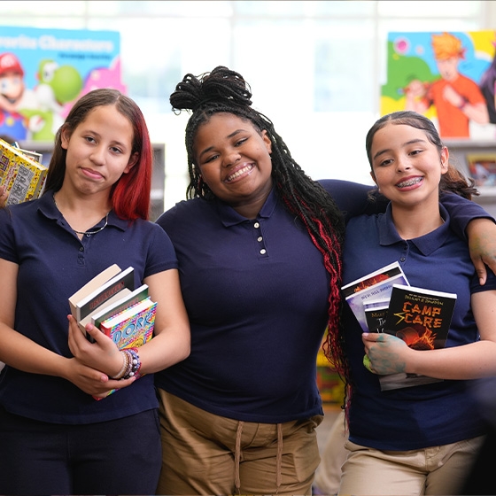 Three students smile in front of Book Fair cases while holding stacks of books