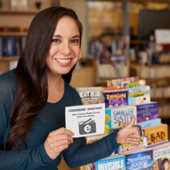 A smiling teacher gives a thumbs up next to a stack of new books from the Book Fair
