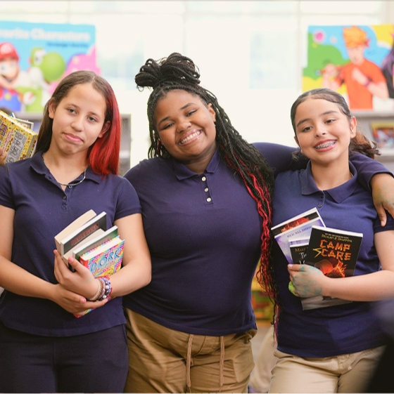 Three students smile in front of Book Fair cases while holding stacks of books