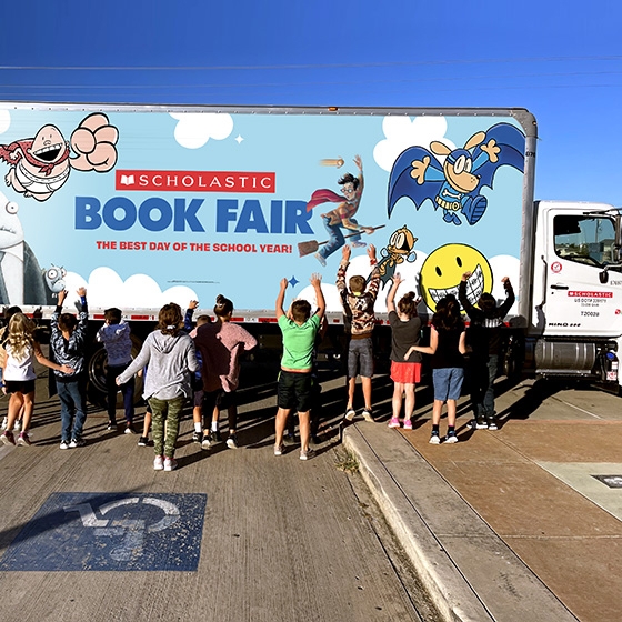 A group of students greet a Book Fair truck excitedly. The truck has the Scholastic logo and text that reads THE BEST DAY OF THE SCHOOL YEAR